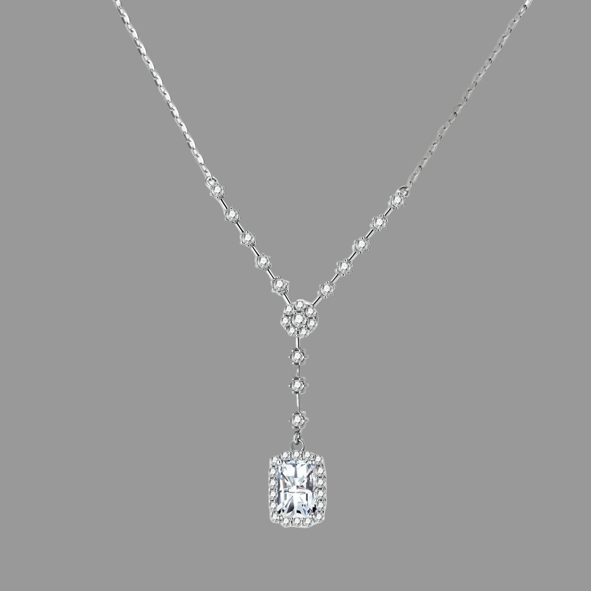 A silver-toned Y-shaped necklace featuring a rectangular emerald-cut clear gemstone pendant surrounded by small clear stones, displayed on a light cream background.