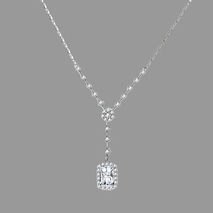 A silver-toned Y-shaped necklace featuring a rectangular emerald-cut clear gemstone pendant surrounded by small clear stones, displayed on a light cream background.