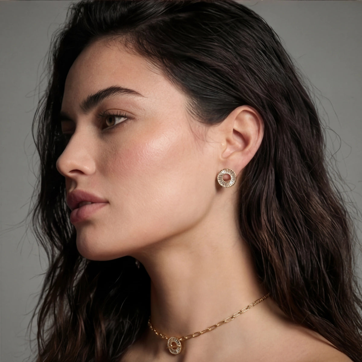 Close-up of a woman wearing gold earrings and necklace against a gray background