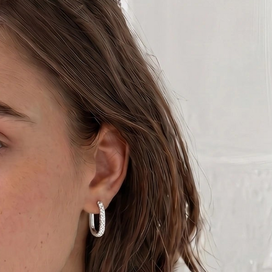 A close-up of a person's ear wearing a small silver hoop earring with a textured pavé-style surface, against a light neutral background.