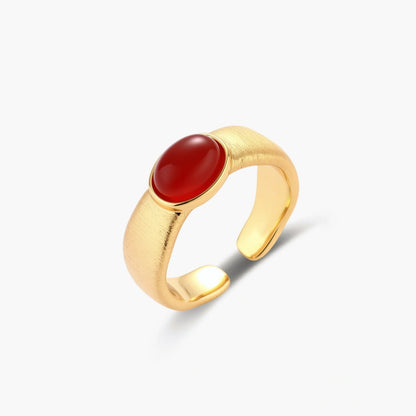 Close-up of brushed gold-plated ring with deep red oval stone, featuring adjustable band design on neutral background.
